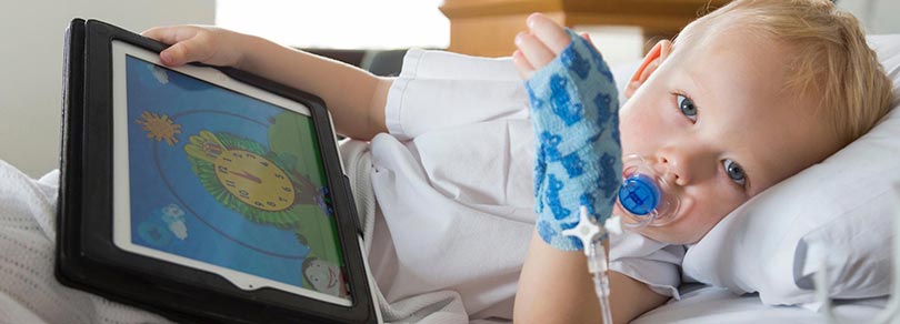 A young boy in a hospital bed holding a digital tablet in one hand and a bandage covering and intravenous line in his other hand, looking at the camera