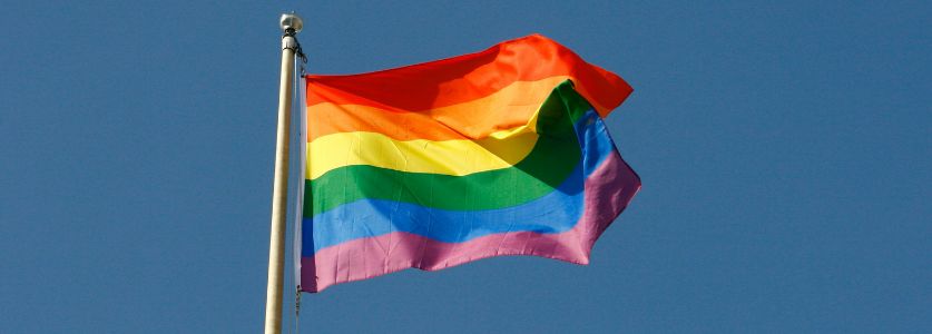 A rainbow flag flaps in the breeze against a clear blue sky.